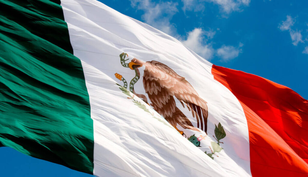 Mexican flag waving against a bright blue sky with scattered clouds.