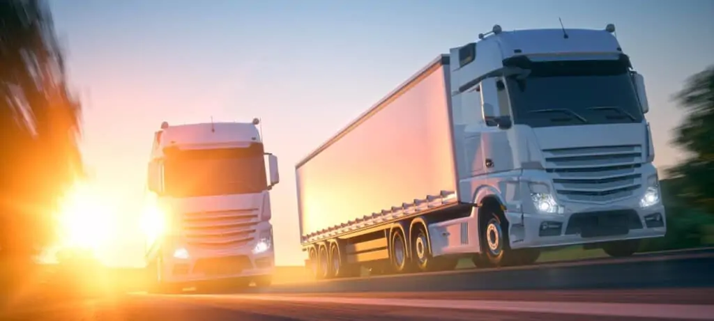 Two white semi-trucks driving toward the camera at sunset with headlights on.