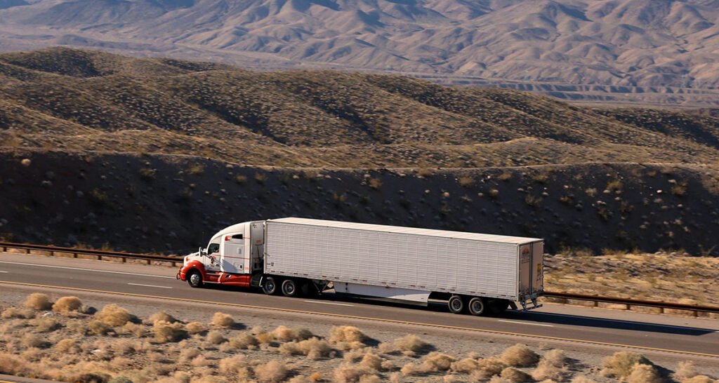 Semi-truck hauling a trailer along a desert highway near rolling hills.