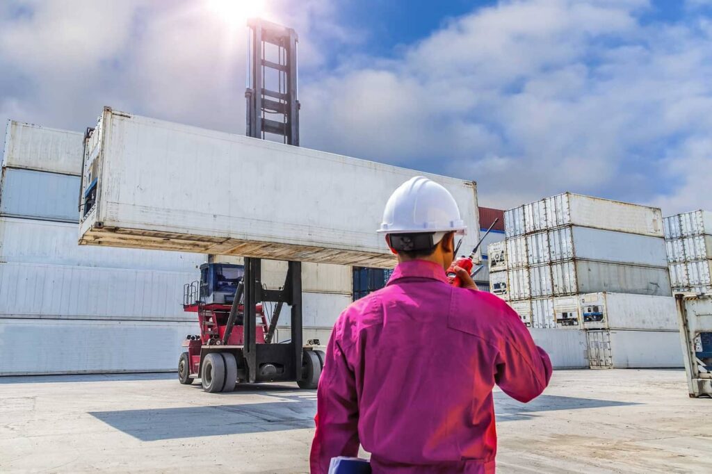 Dockworker in a hard hat directing a forklift lifting a shipping container.