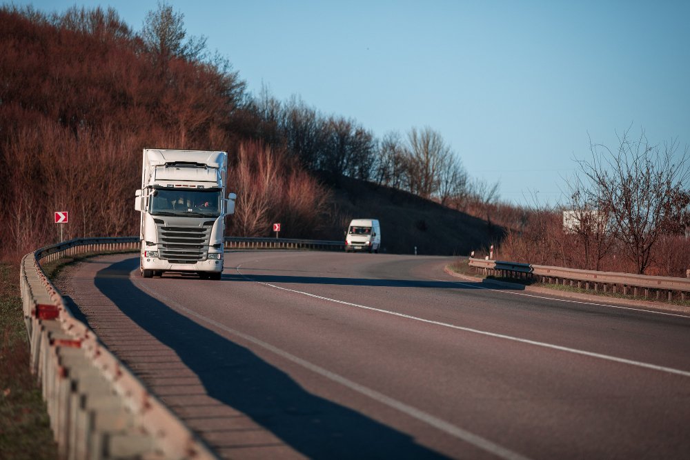 White cargo truck driving on a curving highway with a white van behind it.